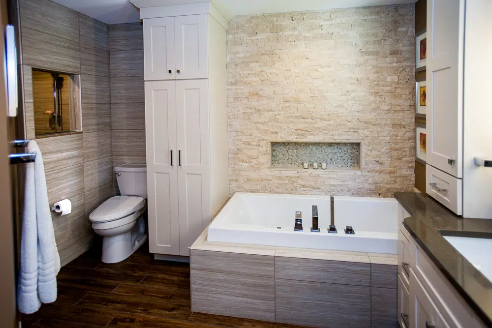 This St. Louis master bathroom is the perfect mix of modern, rustic and traditional. Featuring white cabinetry, quartz countertops, a walk-in shower, porcelain “wood” flooring, mosaic tile backsplash, and a stone wall behind a large and inviting tub.